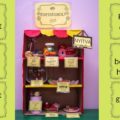kartondobozban berendezett játék édességbolt, mellette a kínálat, zoknifánk, pompon cukorka, szivacs tortaszelet és bonbon