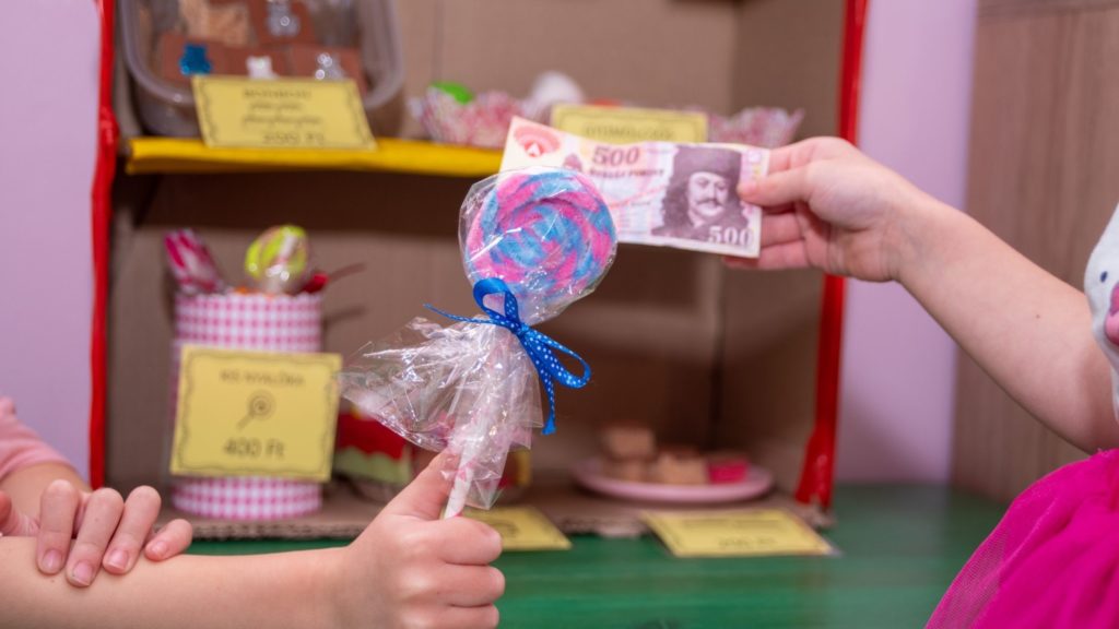 nyalókáért cserébe pénzt nyújt át egyik kislány a másiknak